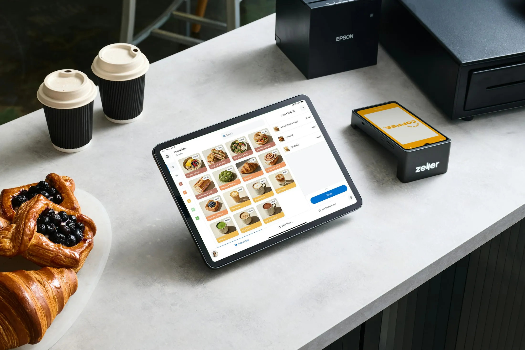 Kassen-Tablet mit Bondrucker und Kassenlade auf einem Tresen, daneben Kaffeebecher und Gebäck